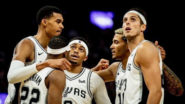 The San Antonio Spurs' starting unit (minus Devin Vassell) stands in a huddle on the court at Frost Bank Center.